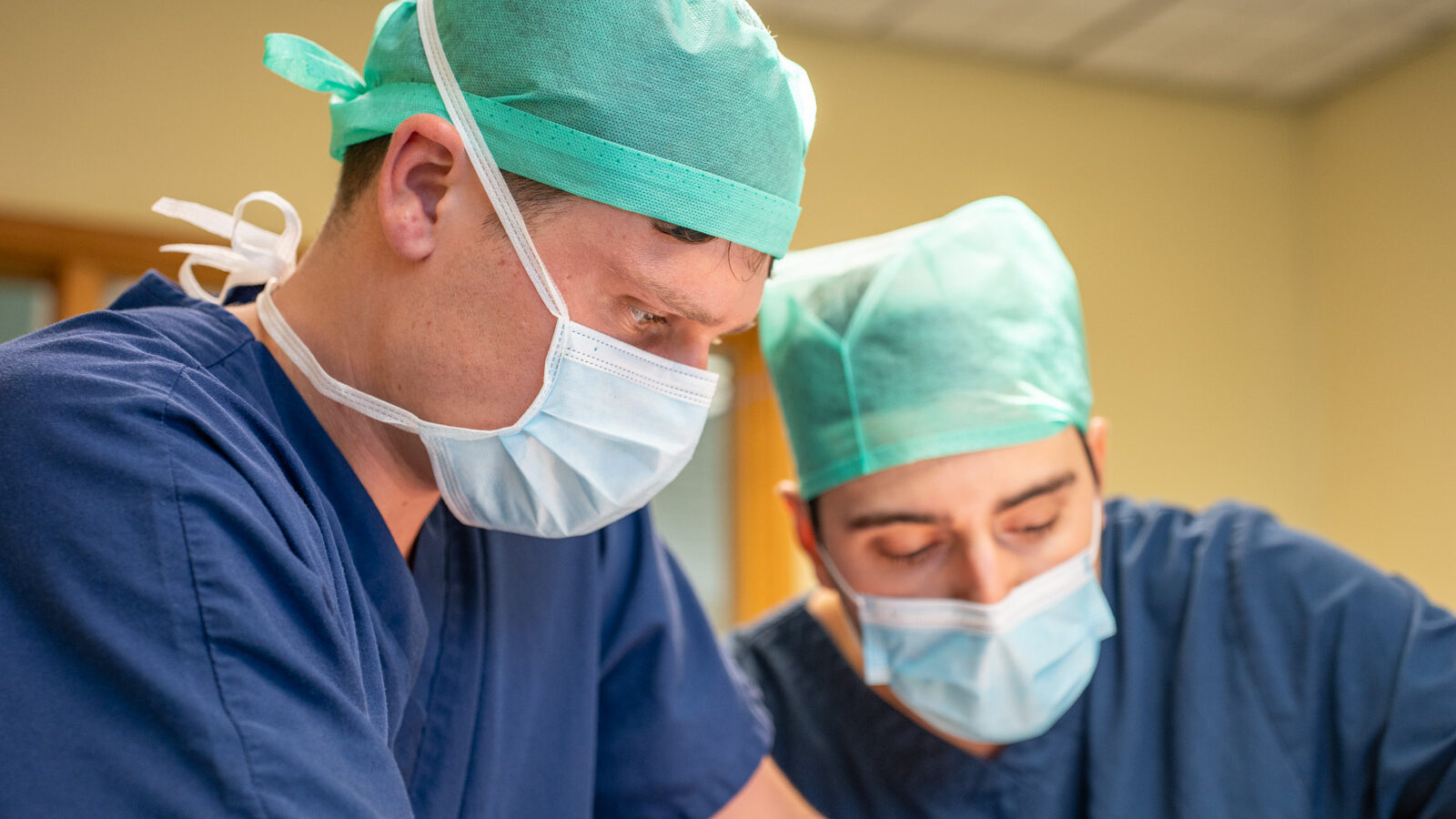 Close up of two surgeons performing surgery. They are wearing green hair nets, white face masks and dark blue scrubs.